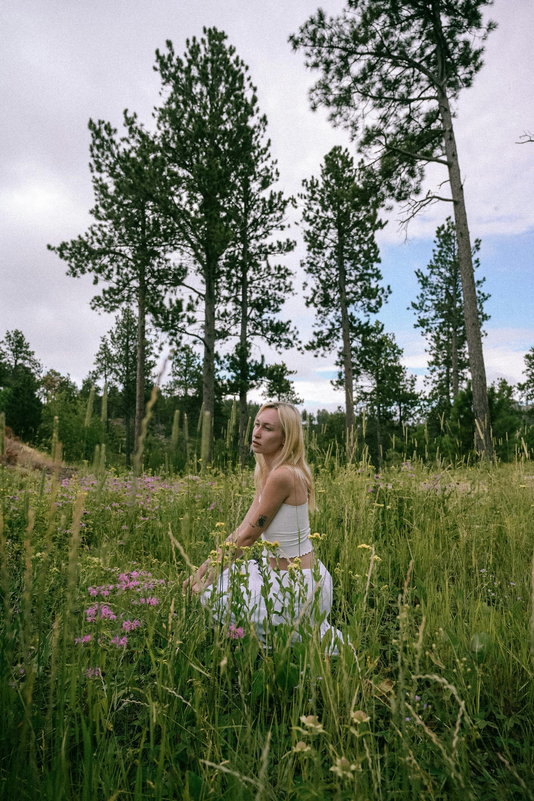 A serene woman seated in a lush meadow with pine trees and wildflowers.