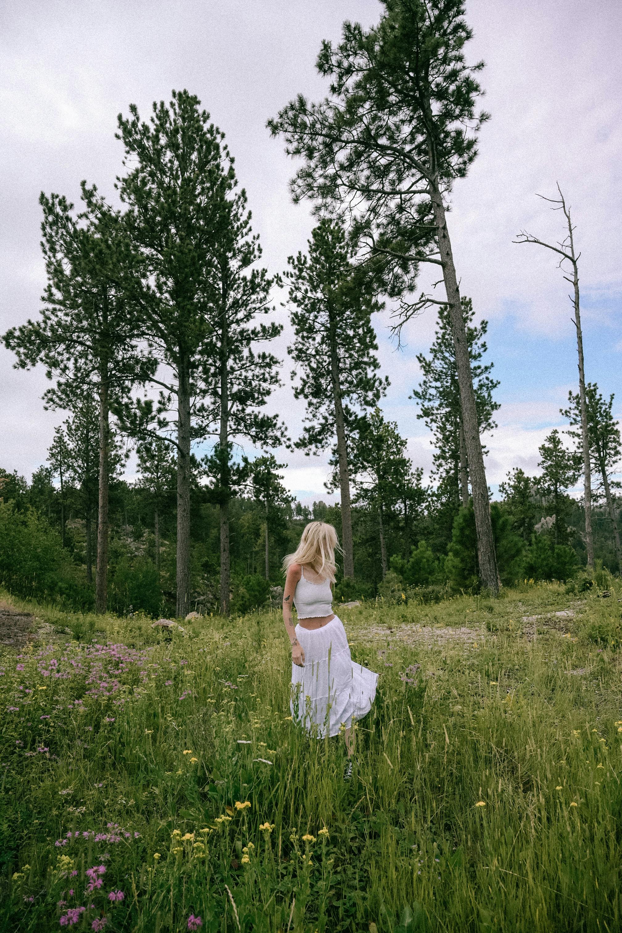 A woman in a white flowing dress standing in a meadow surrounded by tall trees, exuding tranquility.
