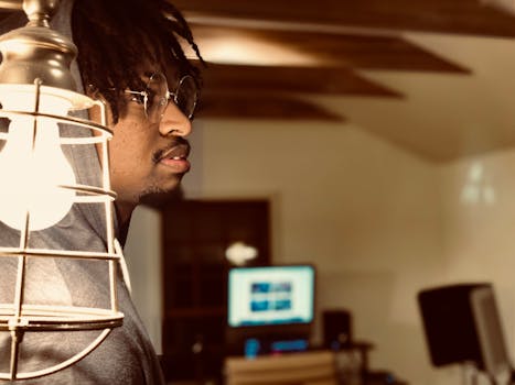 一名非裔美国男子戴着眼镜、穿着连帽衫，站在现代家庭办公室的灯旁，营造出一种舒适、专注的氛围