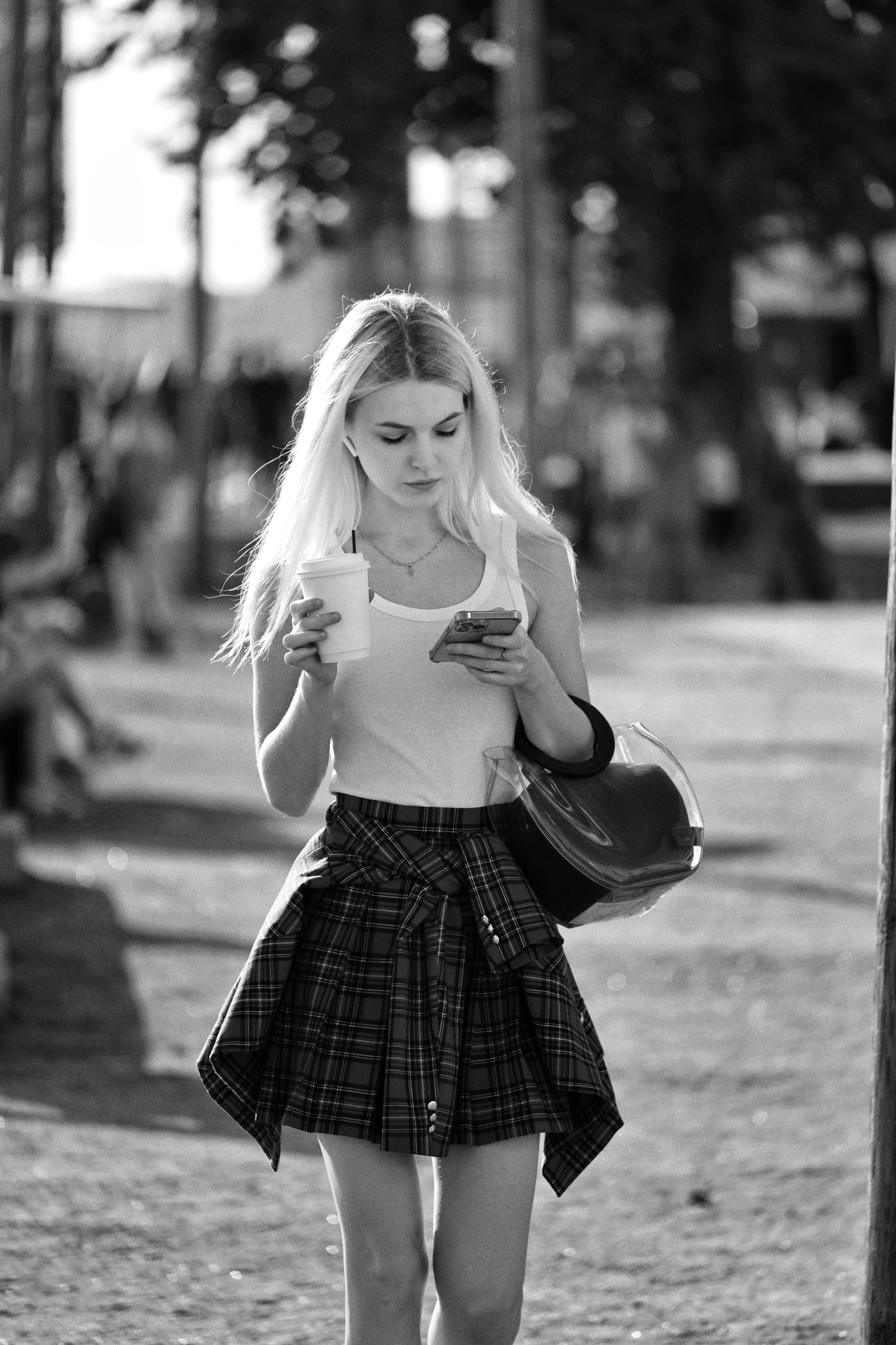 A young woman walks in the park holding a coffee and smartphone during summer.