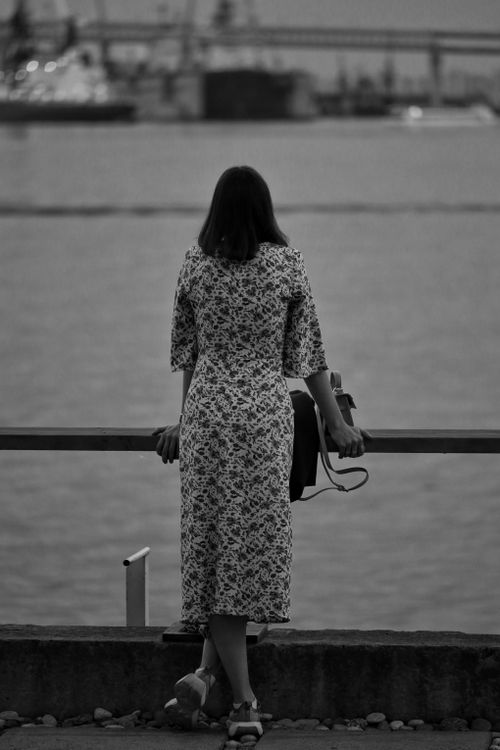 Free Back view of a woman in a floral dress looking out over a waterfront, evoking solitude and contemplation. Stock Photo