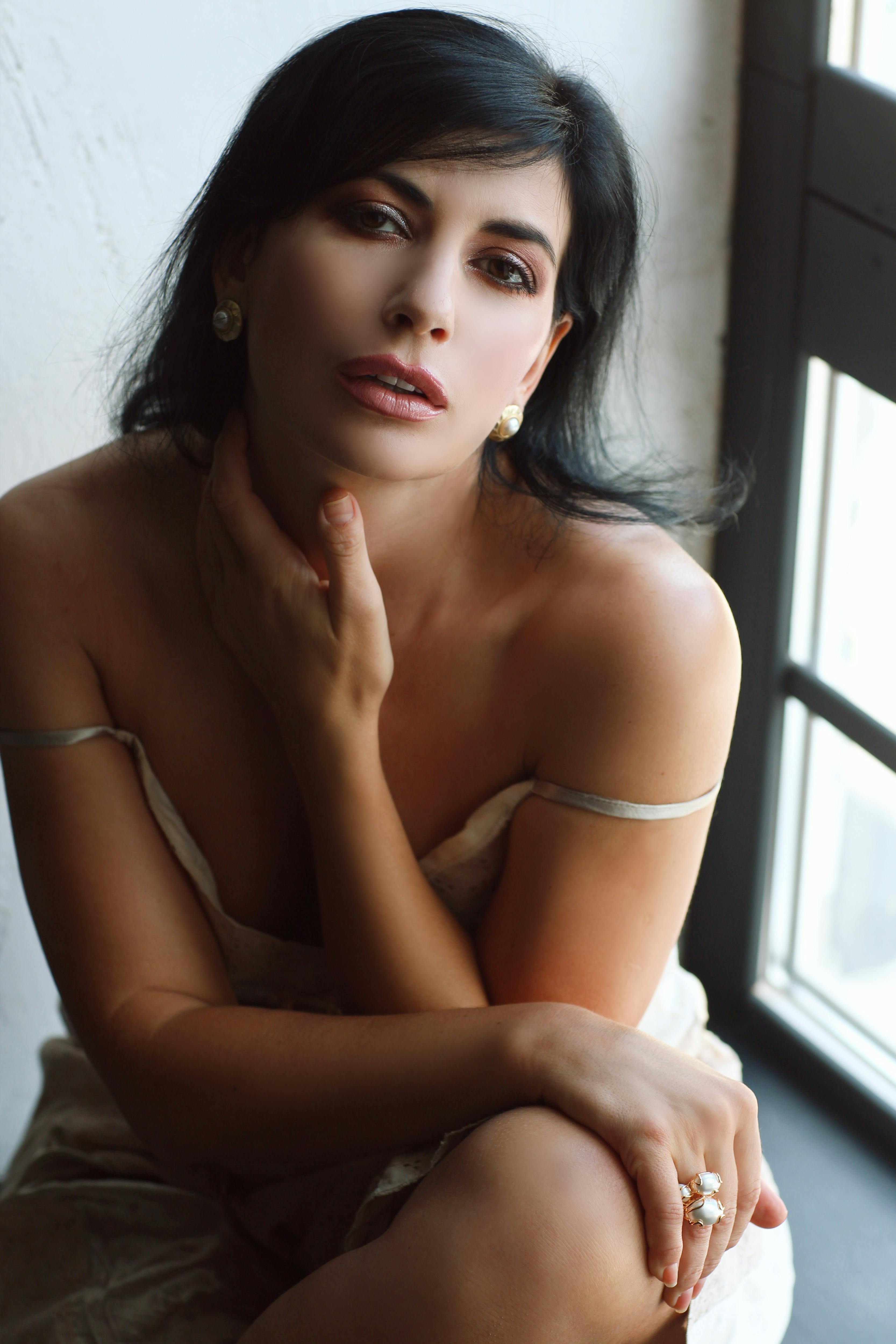 Elegant portrait of a brunette woman with long hair sitting by a window, hand under chin.