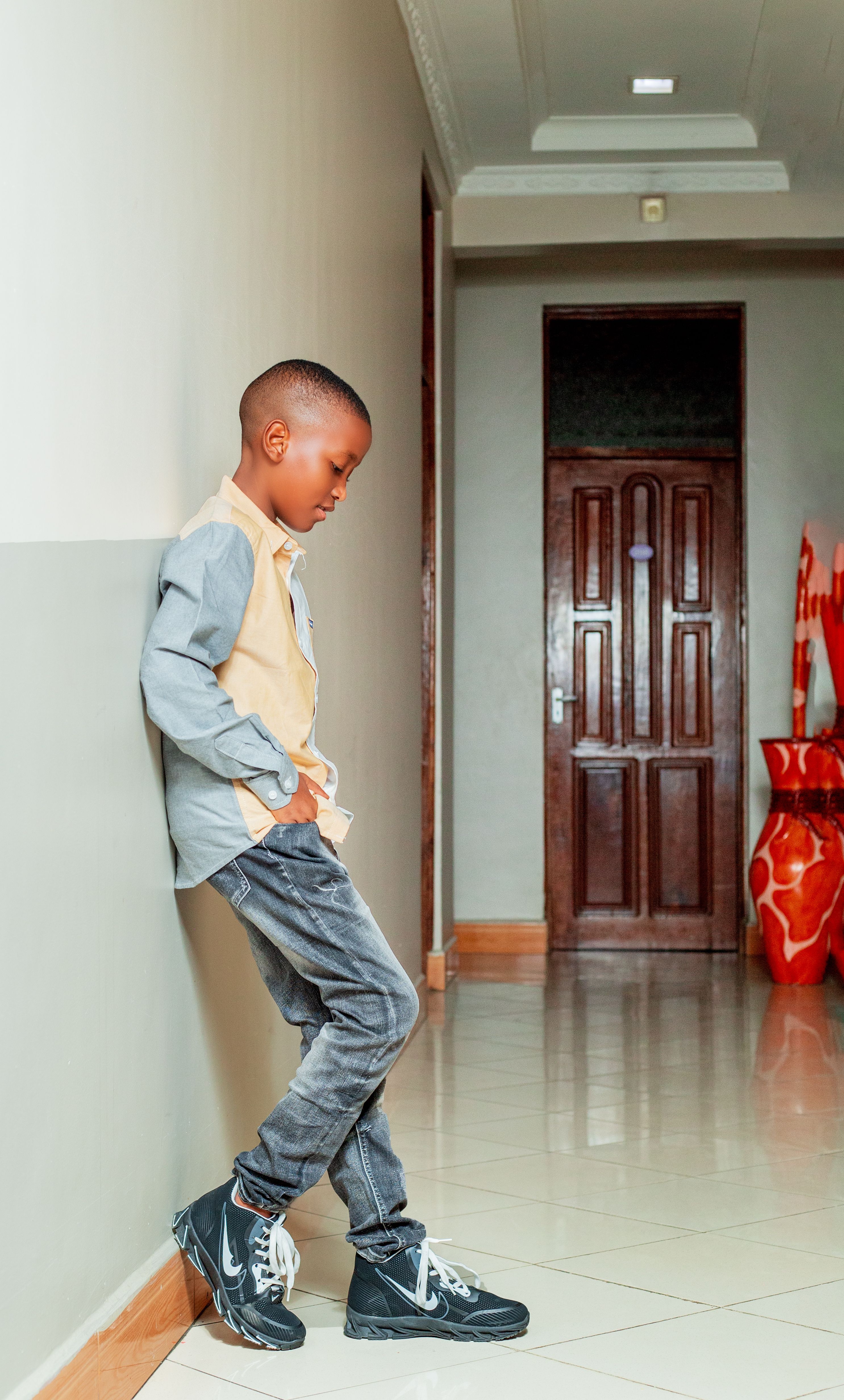 A young boy with short hair stands leaning against a corridor wall, looking down thoughtfully.