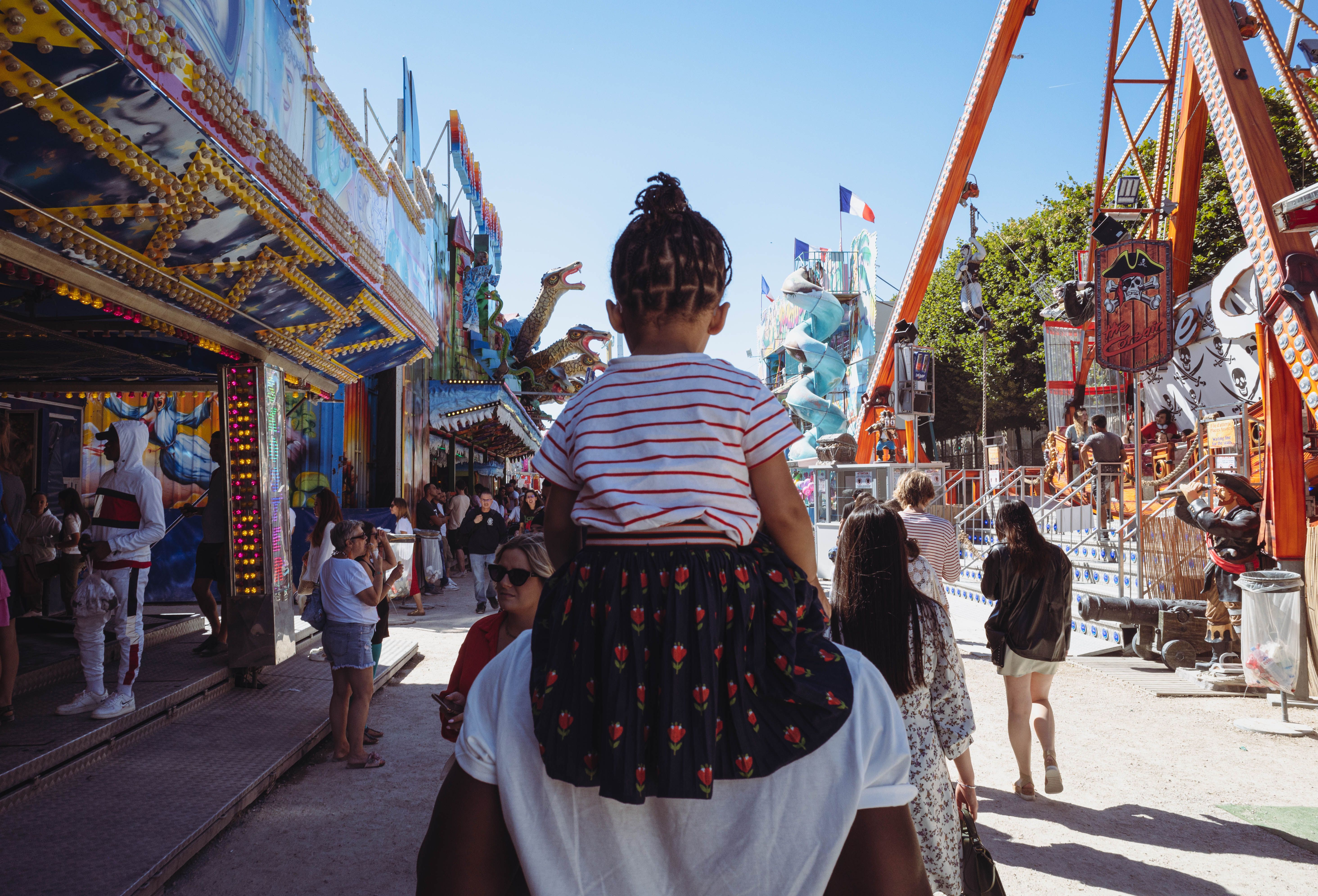 Man carrying his daughter through a bustling amusement park filled with rides under a clear sky.