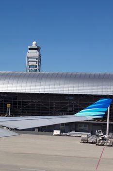 View of airplane wing at modern airport tarmac with terminal and control tower under clear blue sky.