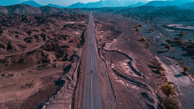 Drone shot capturing a lonely road winding through a vast desert landscape with mountains.