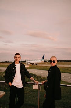 Couple dressed in black and white by airport runway at sunset with airplane in background.