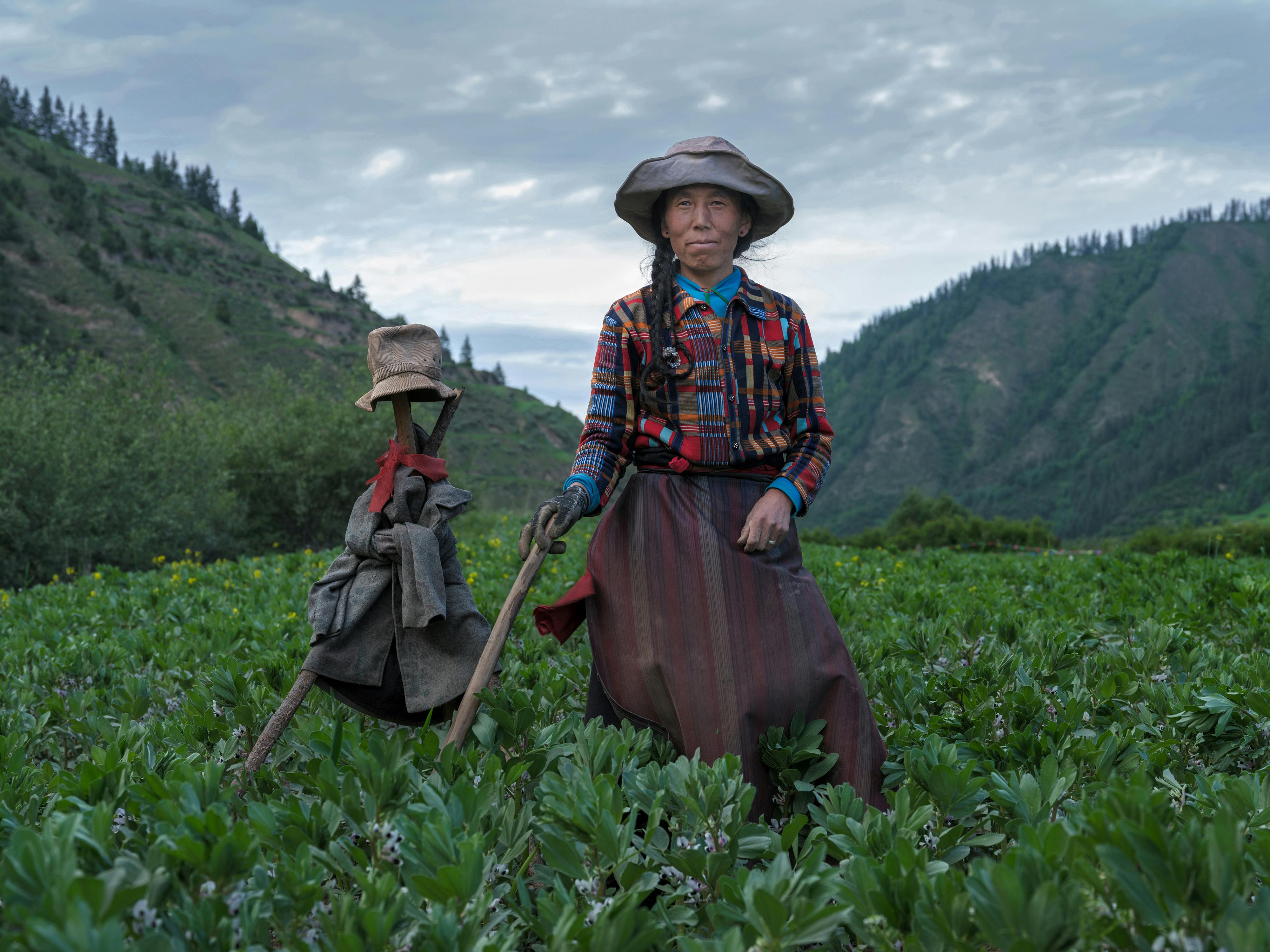 A woman stands in a field beside a scarecrow, showcasing traditional rural life and attire.
