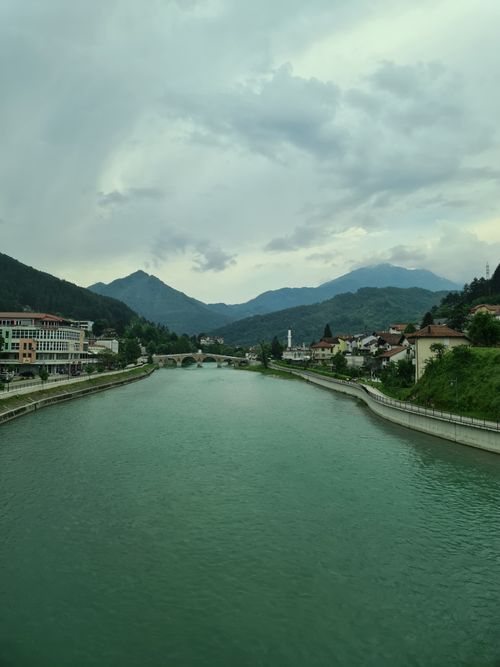 Free Scenic view of a mountain village with a river flowing under a bridge, surrounded by lush greenery and cloudy sky. Stock Photo