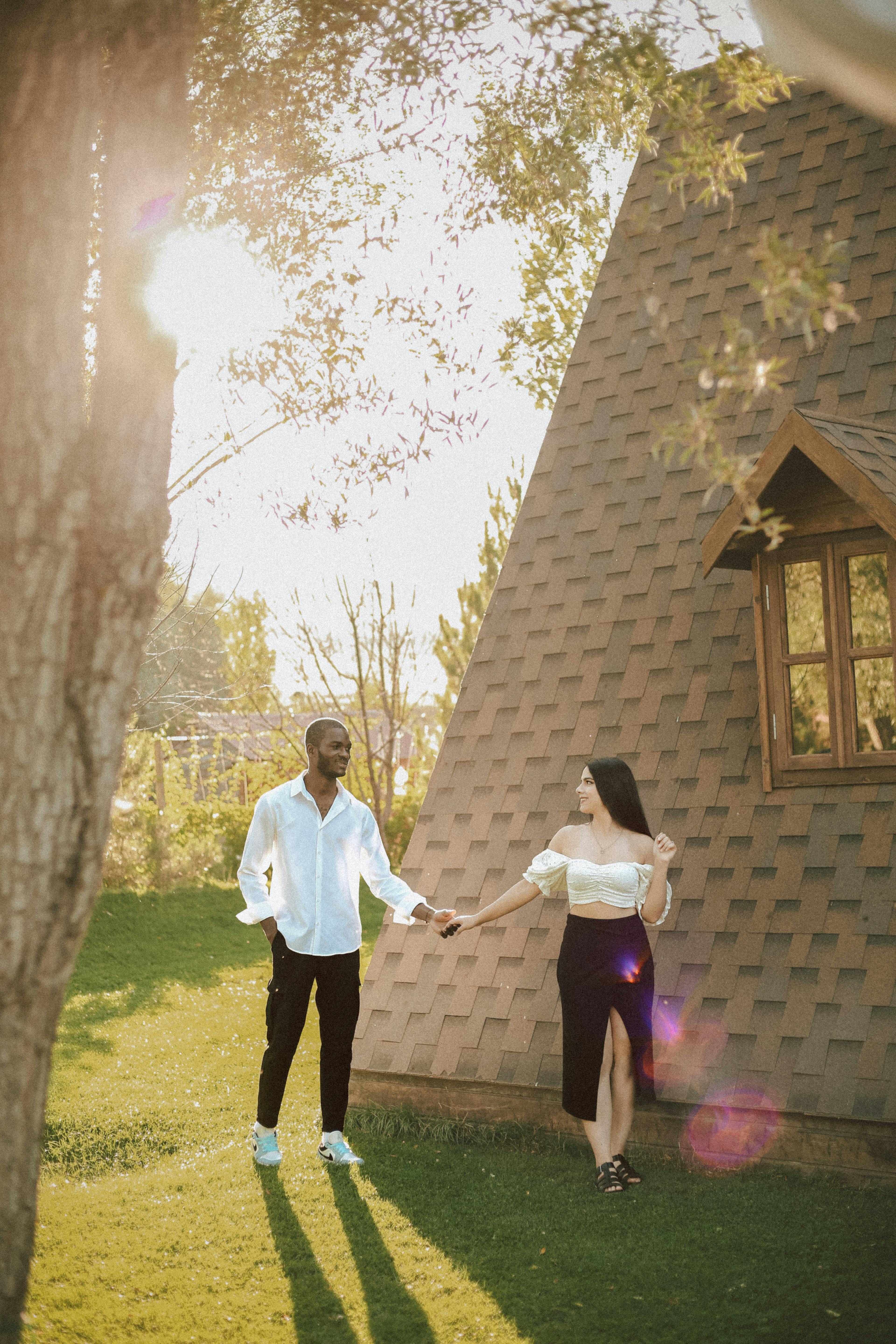 Couple holding hands by a house on a sunny day, with lens flare and lush greenery.