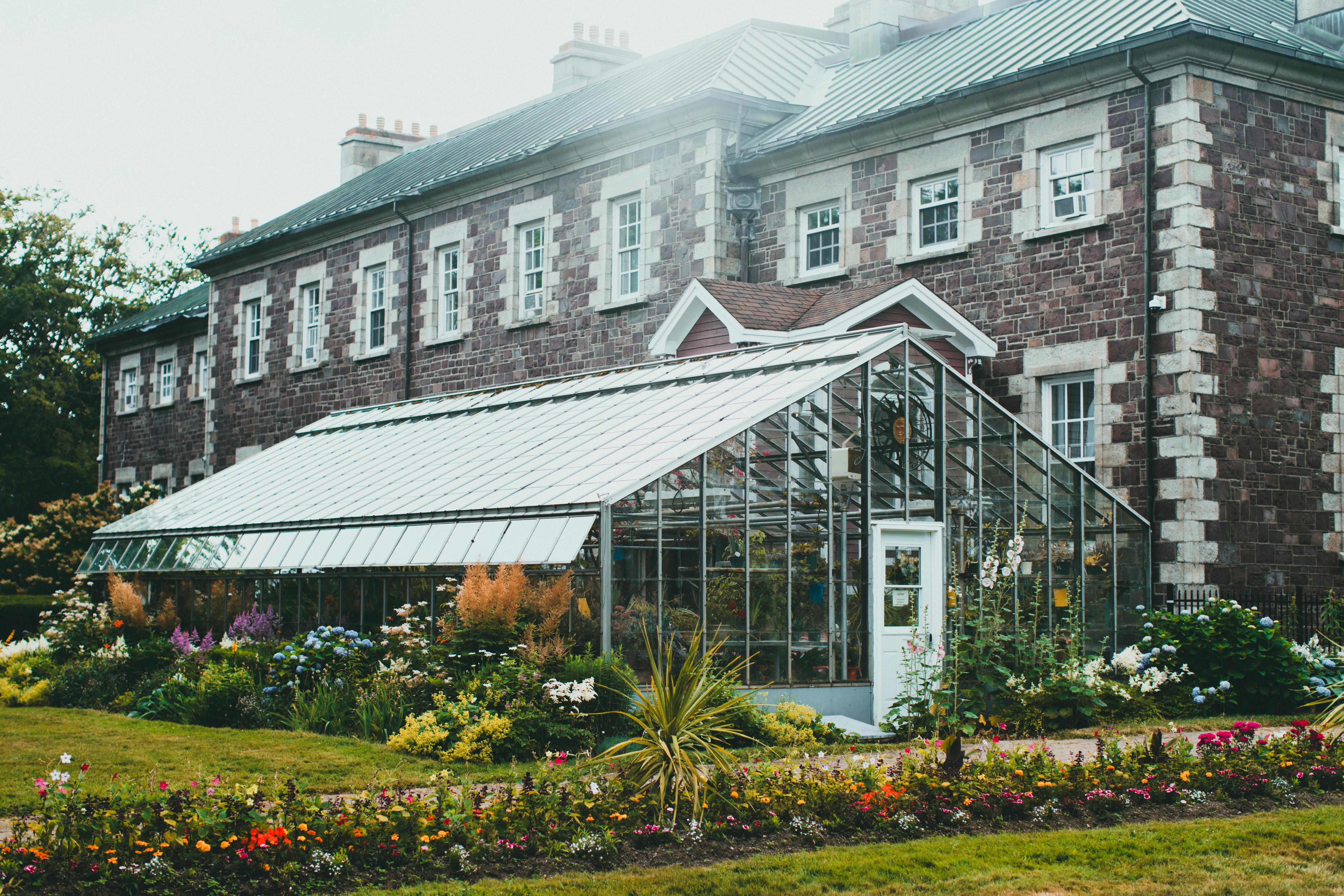 A picturesque greenhouse surrounded by vibrant gardens against a historic stone building.