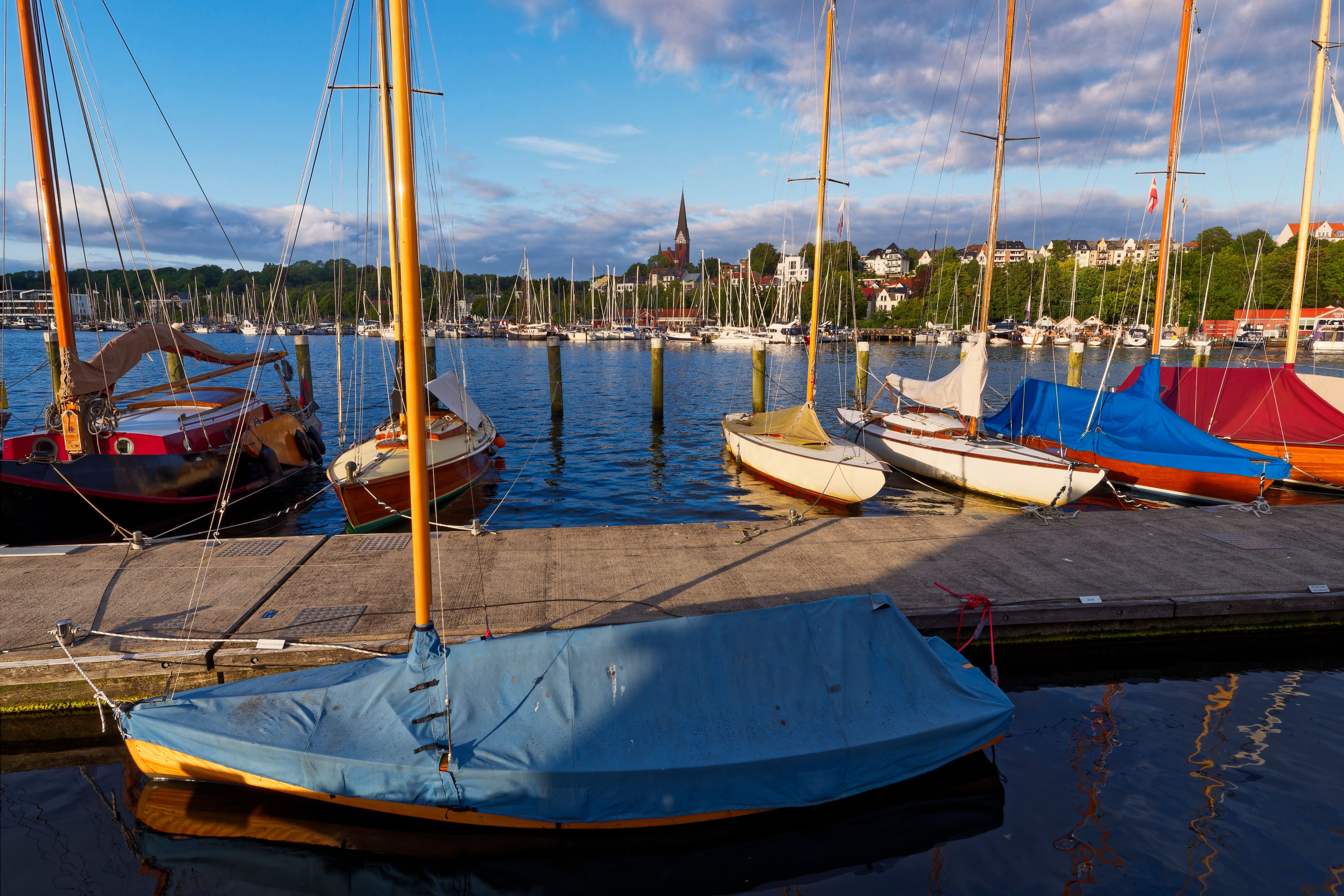 Charming view of sailboats docked by Flensburg harbor with cityscape under summer skies.