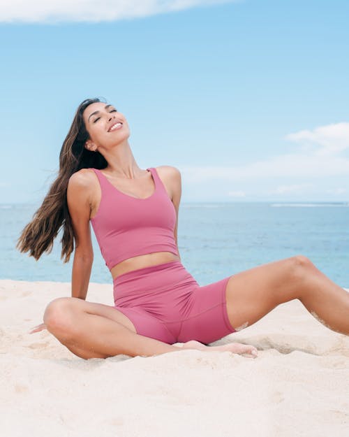 Young Woman in Pink Sportswear Sitting on a Beach and Smiling 