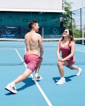 Young man and woman in sportswear stretching on an outdoor tennis court for fitness.