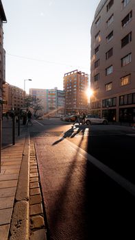 City street at sunset with long shadows and pedestrians crossing under warm sunlight.