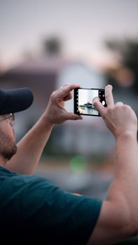Man in cap photographing a scenic view with smartphone outdoors, showcasing modern tourism.