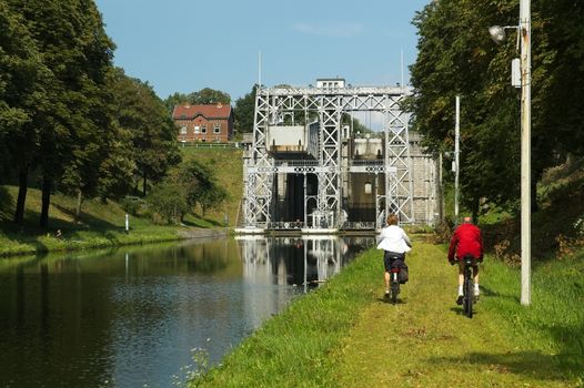 Two cyclists ride along a grassy canal path near a historic boat lift surrounded by trees.