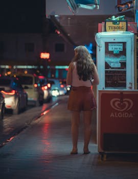 A woman stands on a bustling city street at night, illuminated by neon lights and vehicles.
