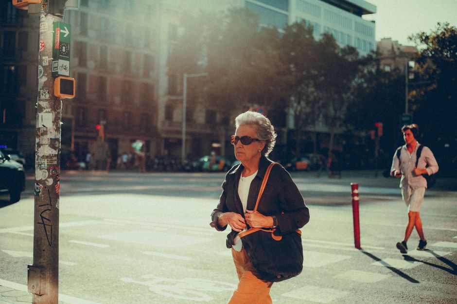 Una mujer mayor con cabello canoso camina en un entorno urbano soleado, encarnando la vida de la ciudad.