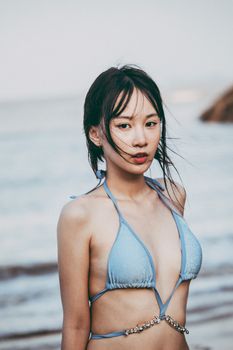 An adult woman with black hair poses in a bikini, standing by the seaside.