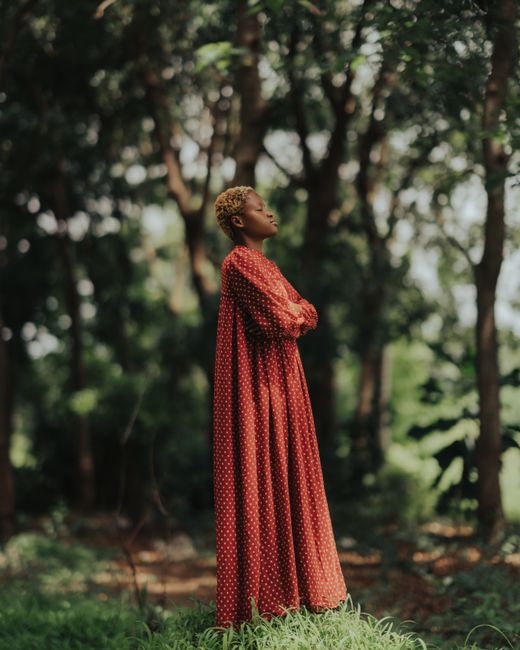 Fashionable woman in a red dress standing in a lush green forest, arms crossed.