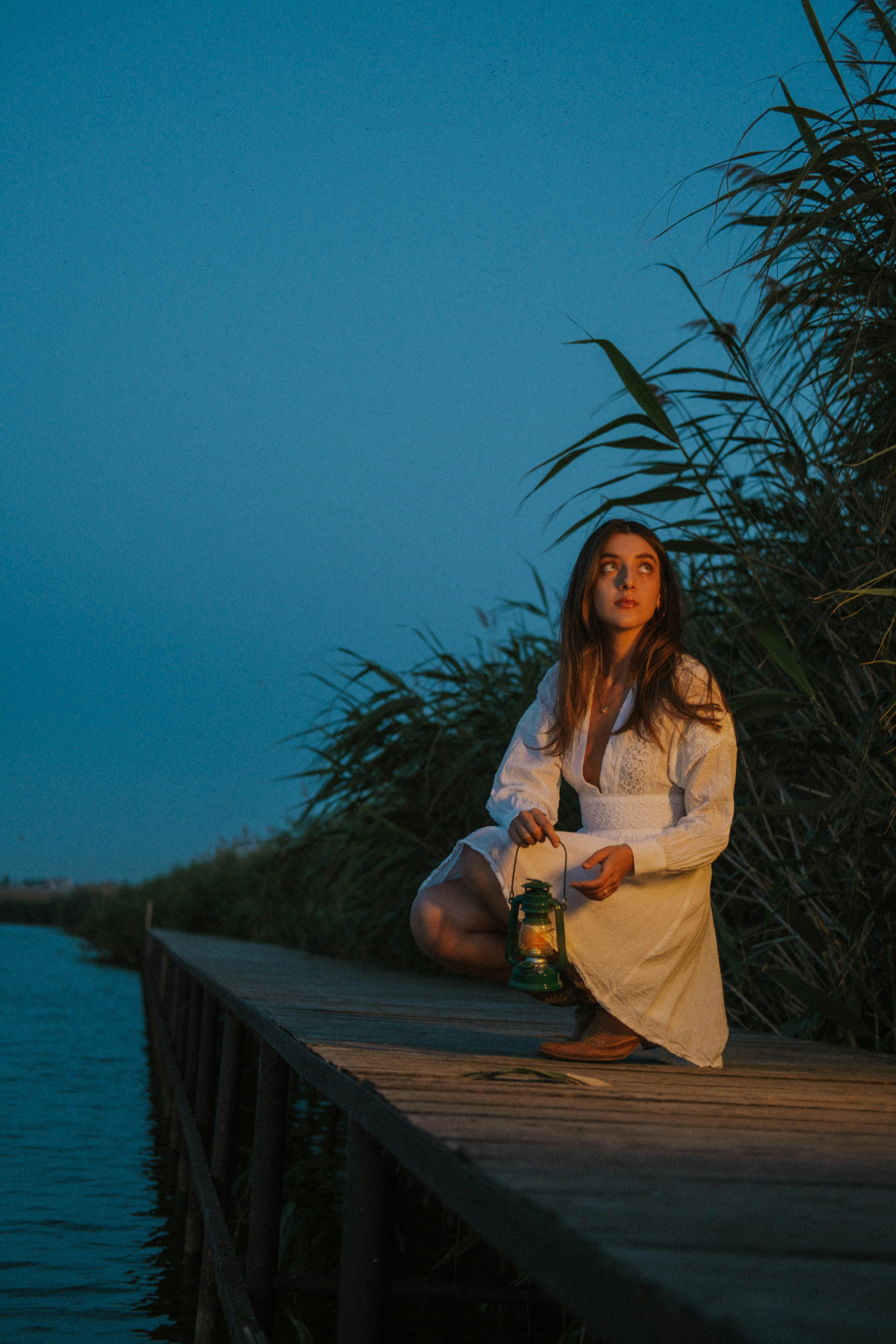 A woman in white kneels on a wooden pier holding a lantern during twilight by a lake.