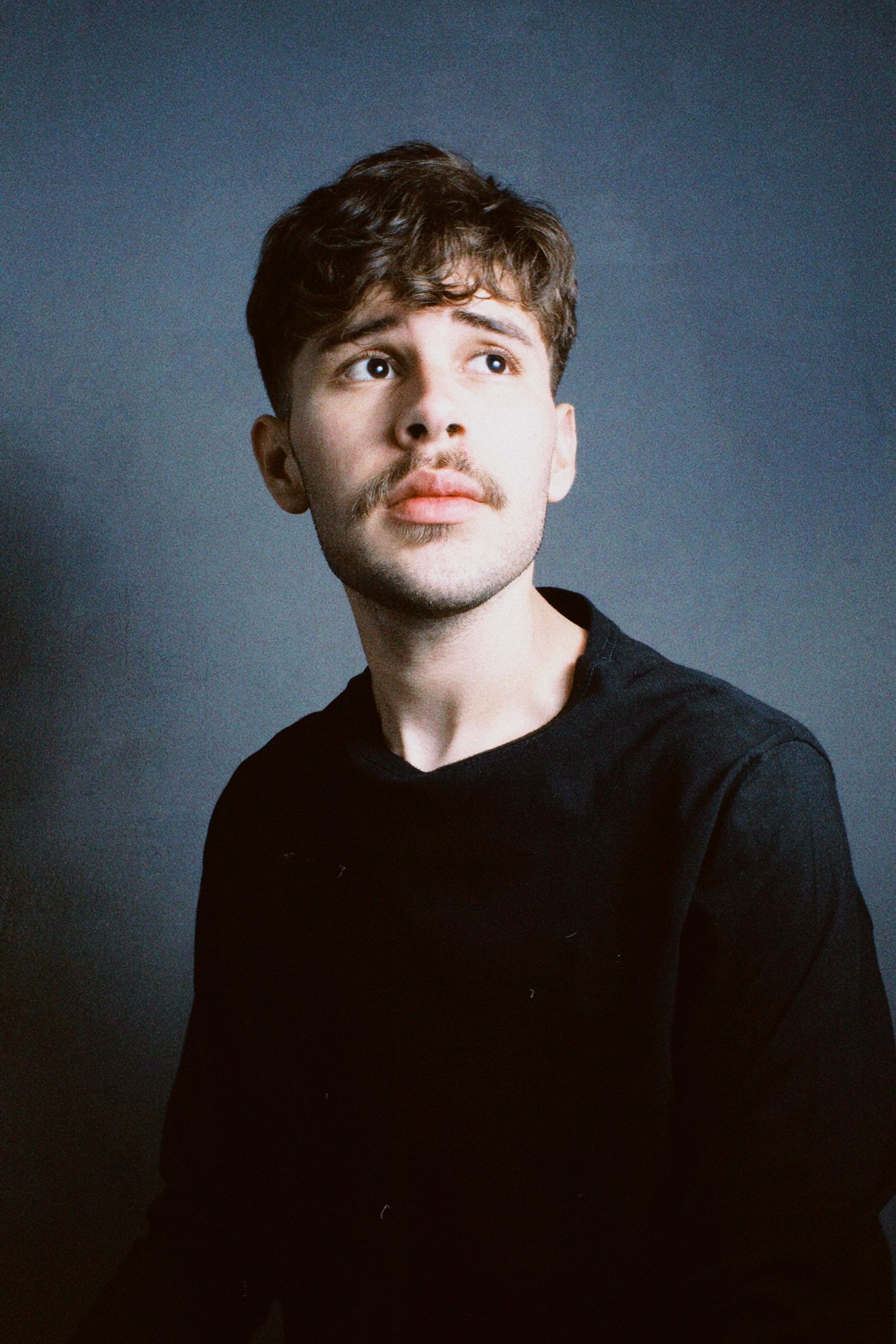 Studio portrait of a young man with a thoughtful expression, featuring a distinctive moustache.