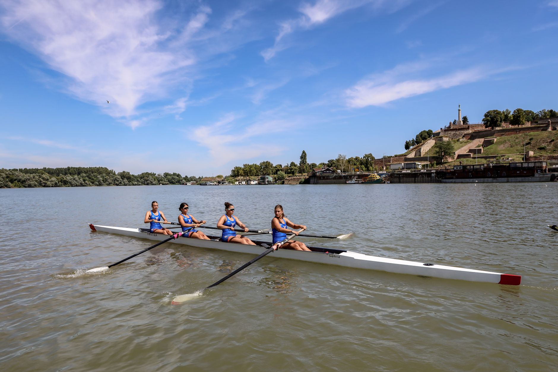 Four female rowers practicing on the Danube River in Belgrade, Serbia with Kalemegdan Fortress in the background.