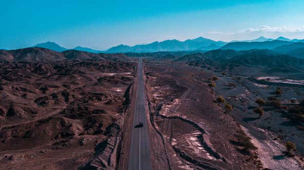 Aerial view of a desert road cutting through a rugged mountain landscape under a clear blue sky.