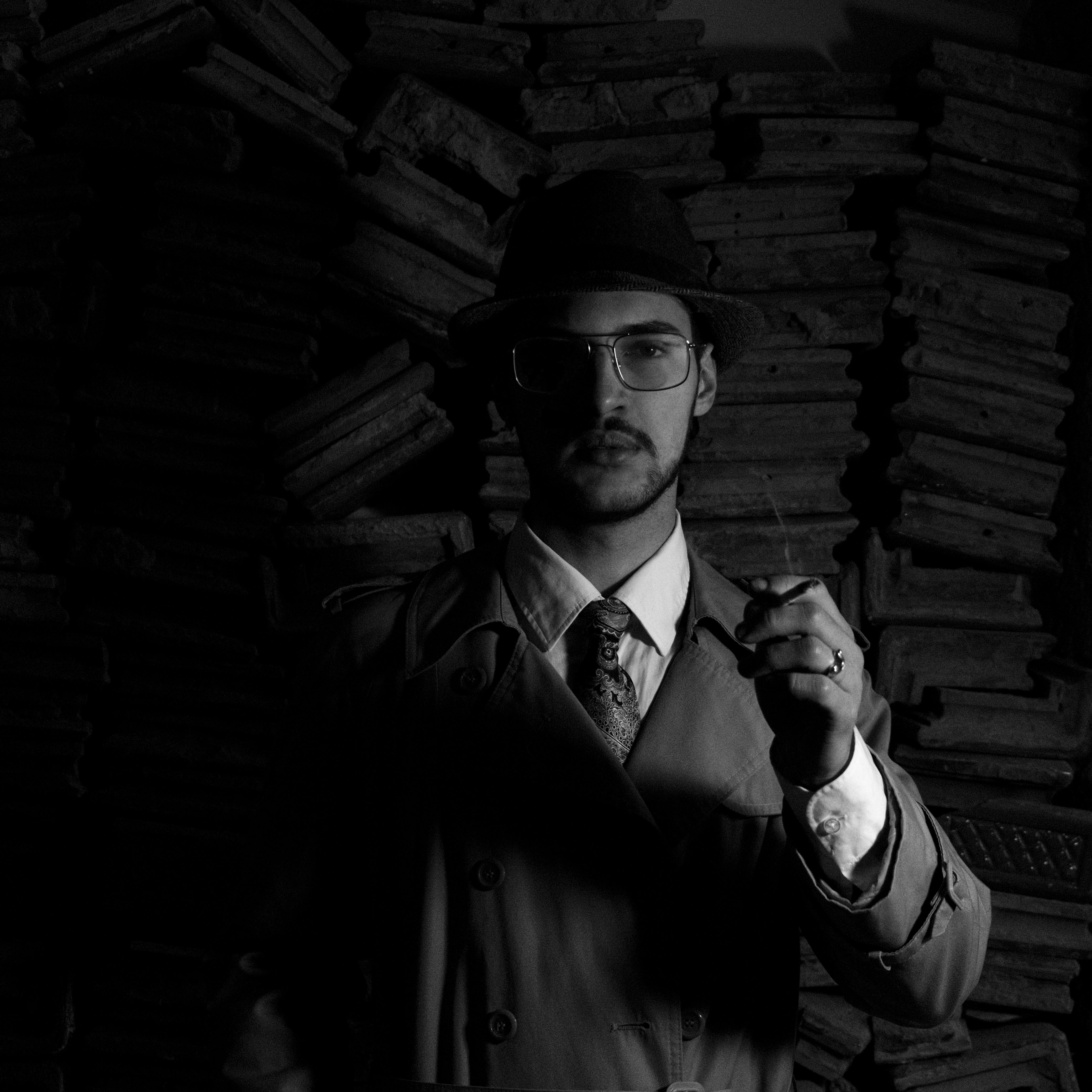 Black and white moody portrait of a man in vintage trench coat and fedora holding a cigarette.