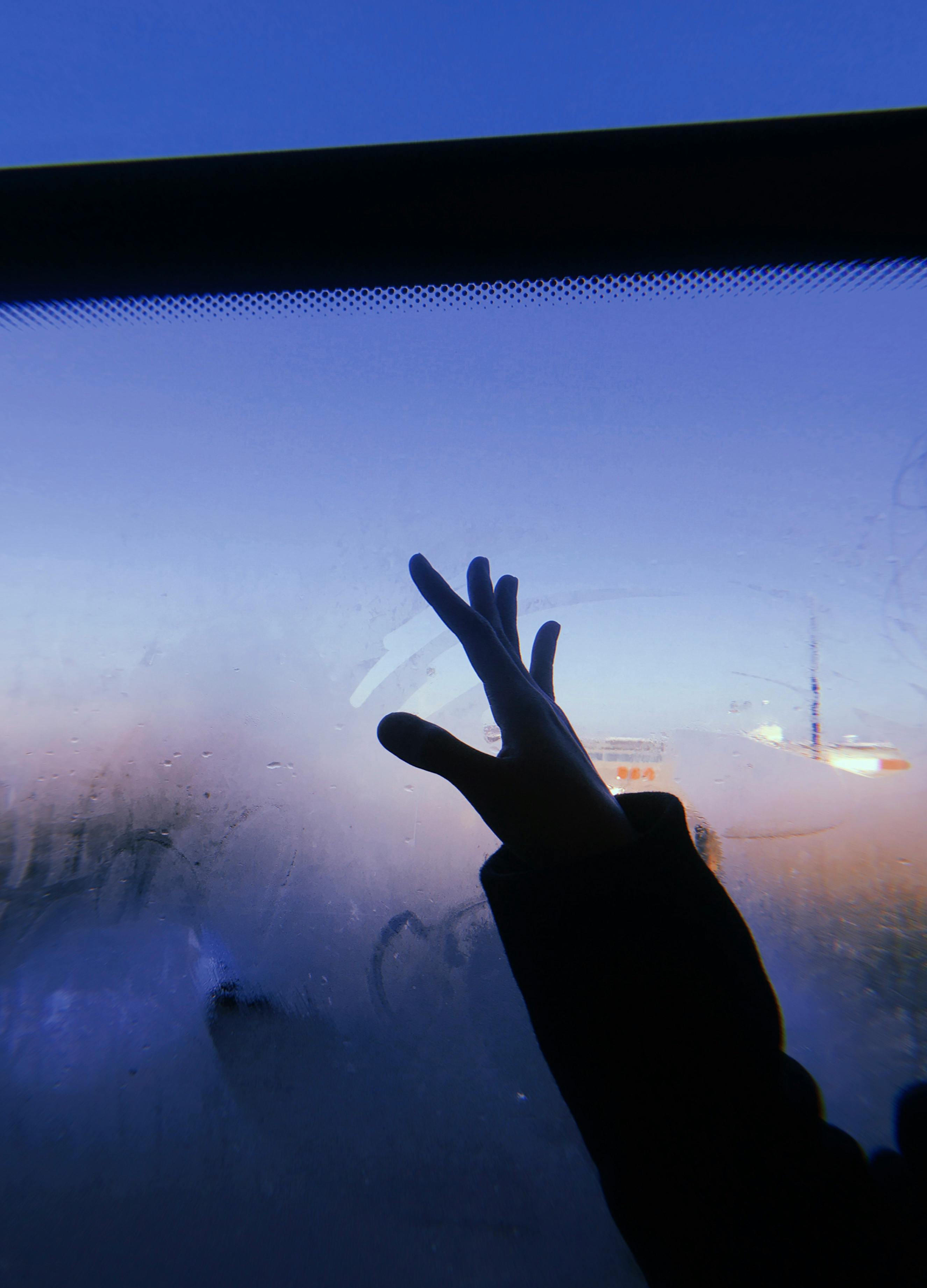 A silhouette of a hand reaches out to a fogged-up window as twilight hues illuminate the sky.