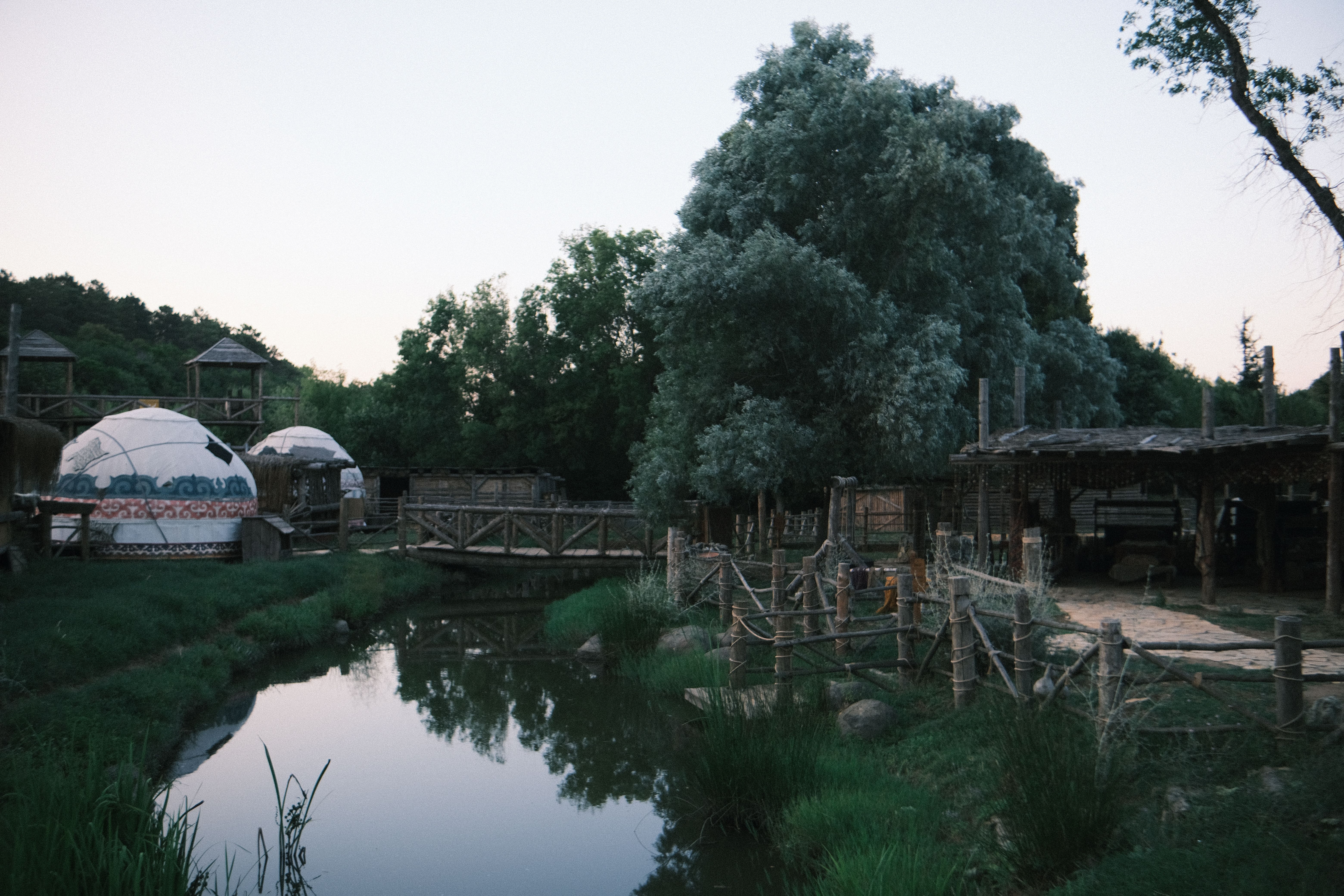 Charming rural scene with yurts, a wooden footbridge, and lush greenery by a tranquil river.