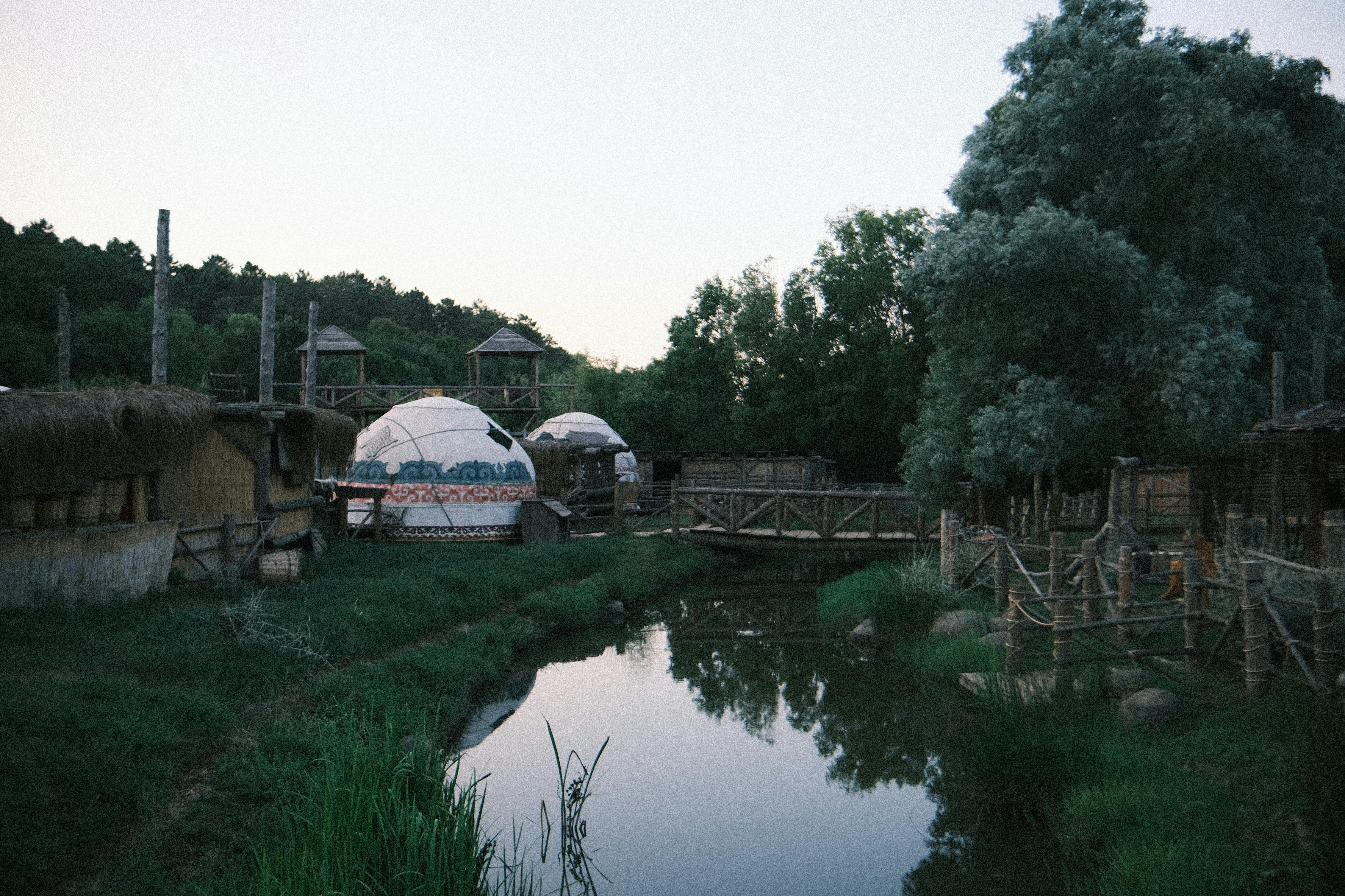 Peaceful village setting with traditional yurts, wooden bridge, and lush greenery by a serene river.