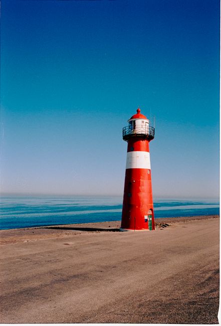 A striking red and white lighthouse stands by the tranquil sea, under a vibrant blue sky.