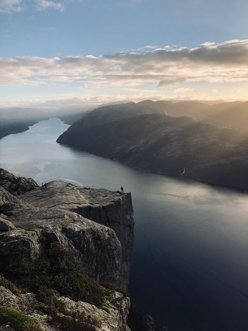 Free A breathtaking view of hikers on a cliff overlooking a fjord at sunrise, capturing the peaceful yet dramatic scenery. Stock Photo