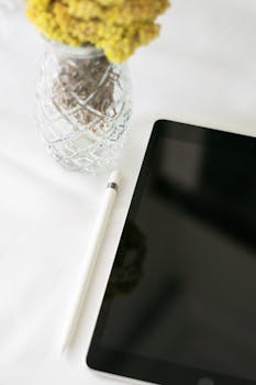 A sleek tablet and pen resting on a white surface beside a decorative vase.