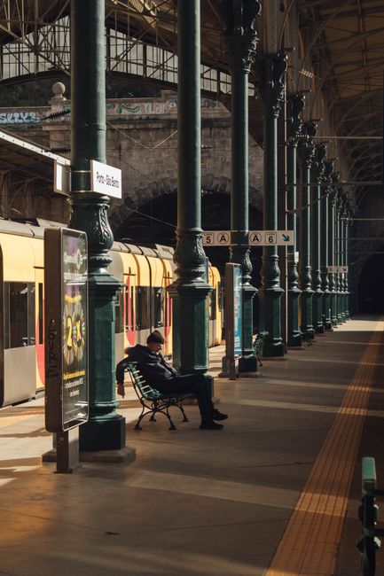 A passenger seated at Porto railway station, Portugal, amidst architectural columns and train, evokes urban travel vibes. — Estação, Portugal.