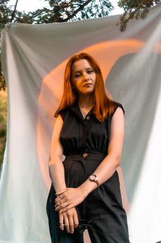 Stylish young woman with red hair posing elegantly outdoors with a backdrop.