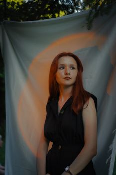 A stylish redheaded woman poses outdoors in a black romper against a soft, artistic backdrop.