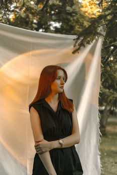 A woman in a black dress stands outdoors, illuminated by soft natural light against a white curtain backdrop.