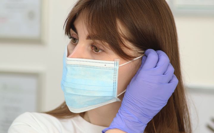Woman healthcare worker wearing gloves and face mask indoors, adjusting PPE for safety.