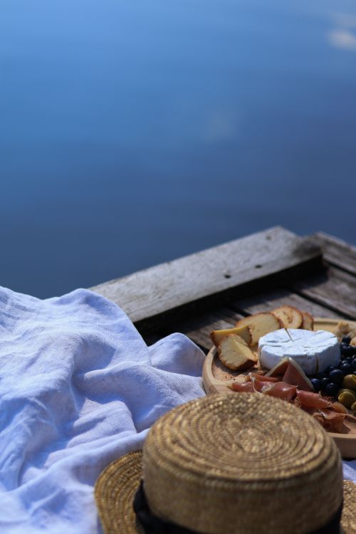 Free A serene lakeside picnic featuring a straw hat, cheese platter, and summer ambiance. Stock Photo