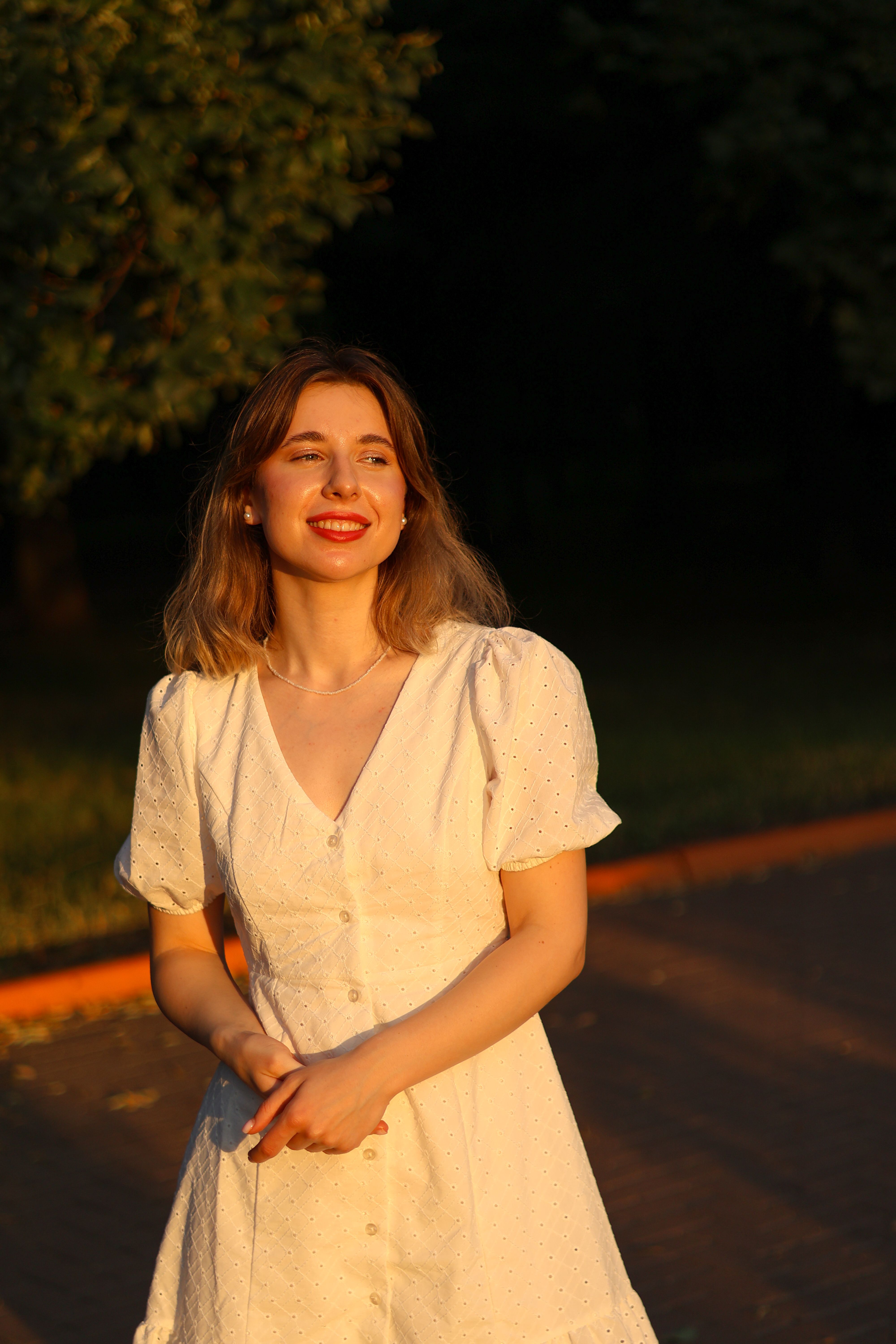 Young woman in white dress smiling warmly during sunrise in a park setting.