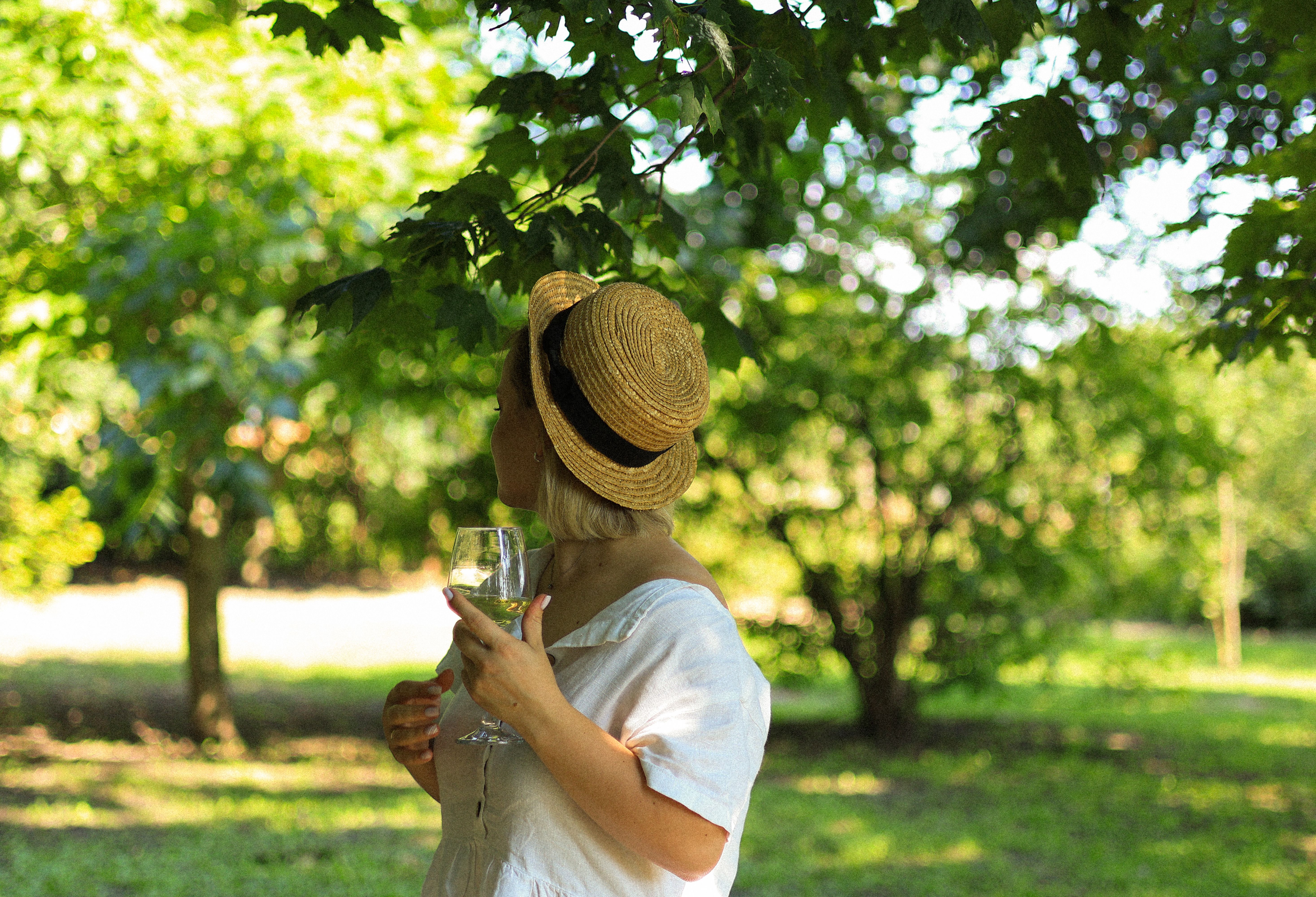A woman in a straw hat holding a wineglass, enjoying a sunny day in a green park.