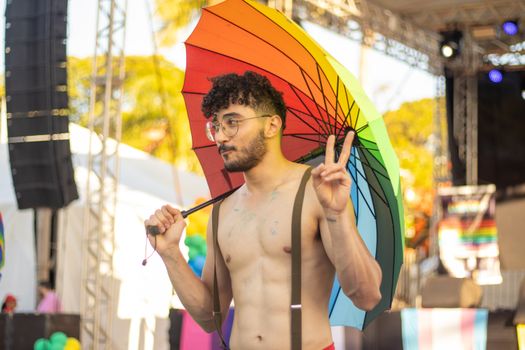 Shirtless man with a rainbow umbrella at a pride event, posing with a peace sign.