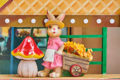 Free Charming Easter bunny figurine with a cart of yellow flowers beside a red mushroom. Stock Photo
