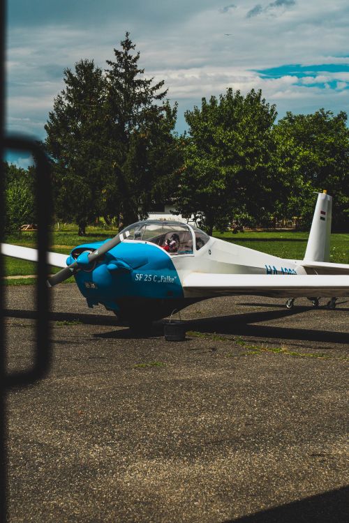 Free Scheibe Falke motor glider parked on tarmac, surrounded by trees in Szombathely, Hungary. Stock Photo