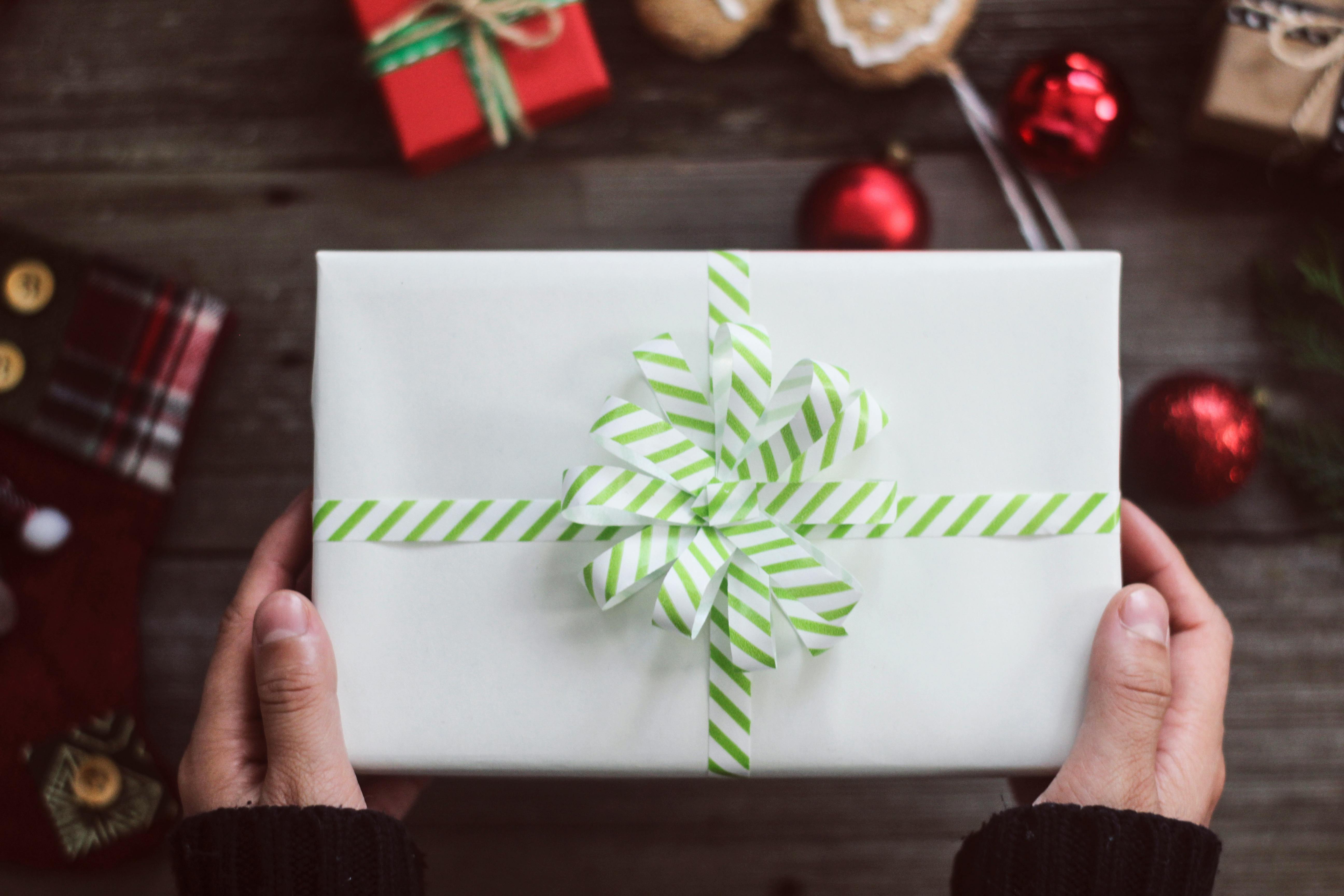 A festive Christmas gift with a green striped ribbon held by hands, surrounded by holiday decorations.