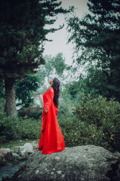 Free Elegant woman in a red gown standing on a rock in a lush park setting. Stock Photo