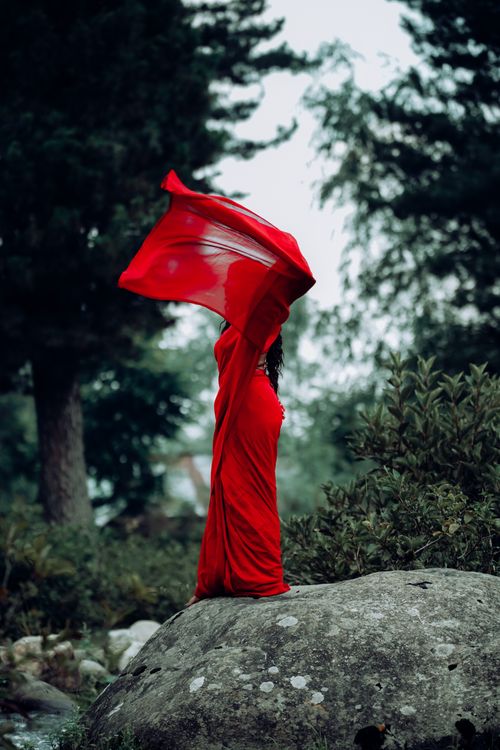 Free Mysterious woman in a flowing red gown standing gracefully on a rock in a lush forest. Stock Photo